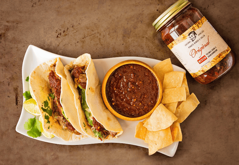 Jar of Pirate Ringo's Salsa with plate of chips and salsa and tacos with salsa.