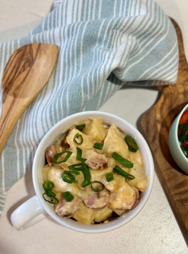 Top view of cheesy sausage and potatoes in a soup bowl topped with scallions, with cutting board, garnishes and tea towel in background.