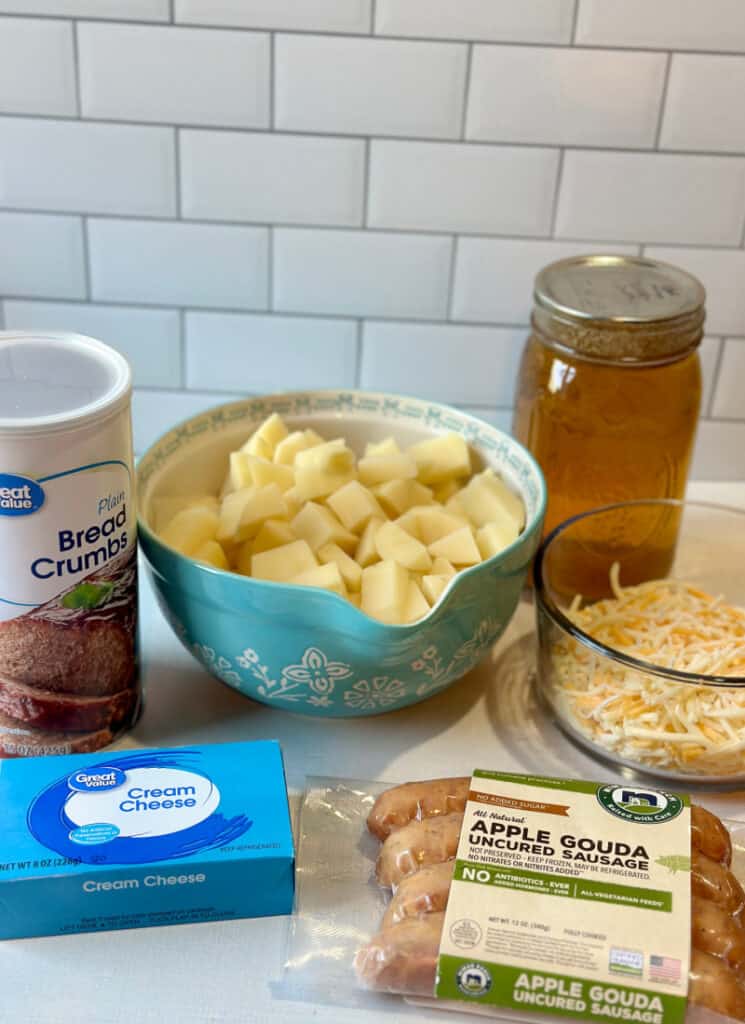 Ingredients on white counter top: potatoes, bread crumbs, chicken broth, shredded cheese, cream cheese, sausage.