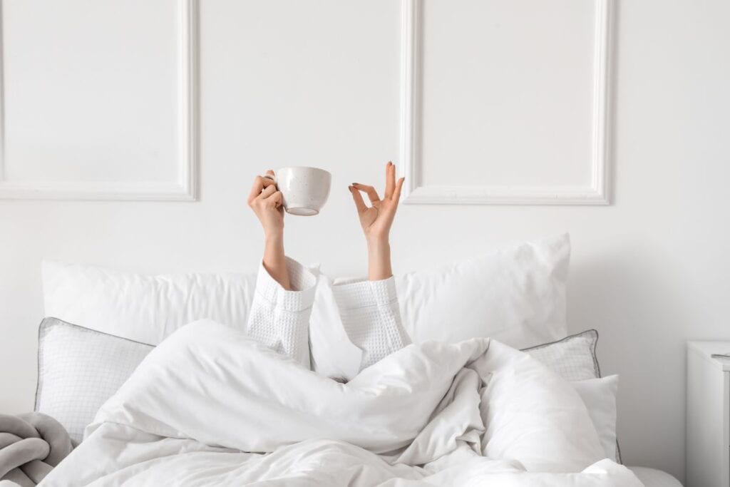Woman under the covers in bed holding up a cup of coffee above her.