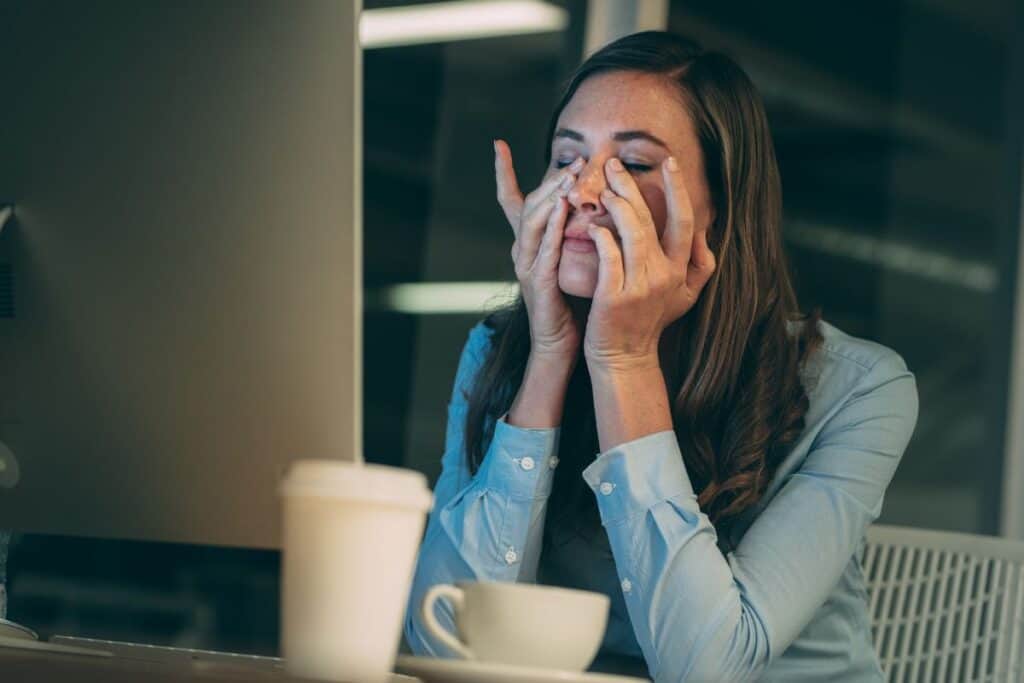 Woman sitting at the computer, rubbing her eyes with coffee on table.