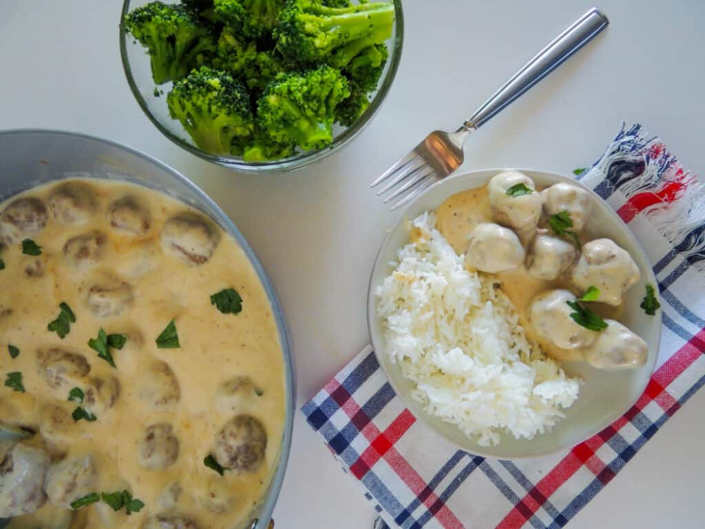 Swedish meatballs on plate with rice and brocolli on the side and skillet with meatballs on table.