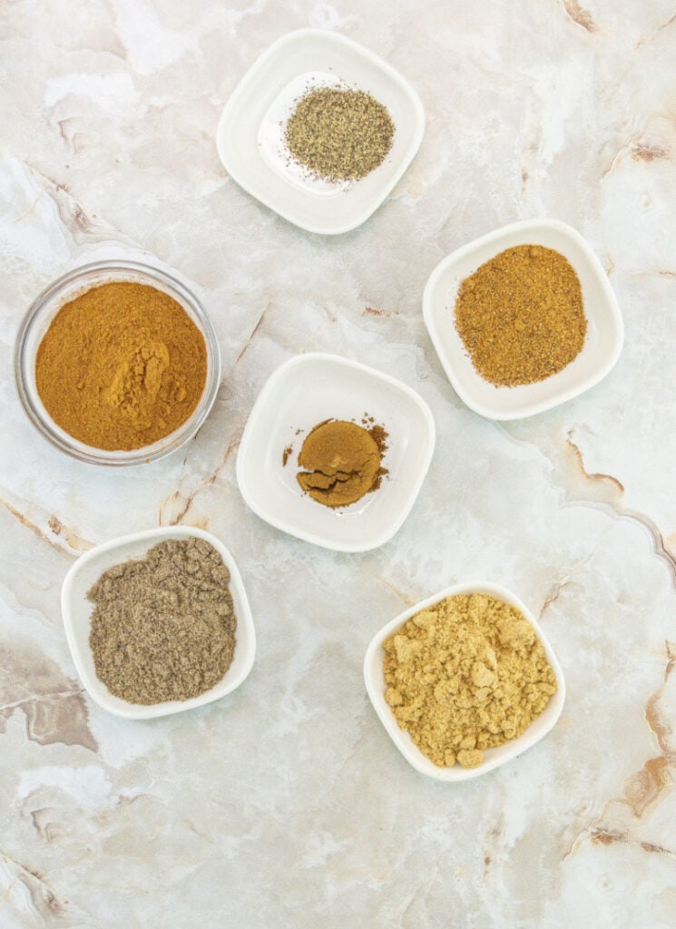 Five small white ceramic dishes with cinnamon, ginger, pepper, cloves, cardamom and nutmeg.