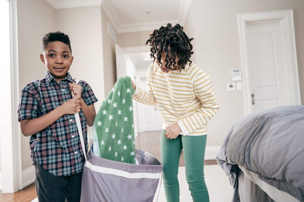 Two kids cleaning up a bedroom and putting laundry into a hamper.