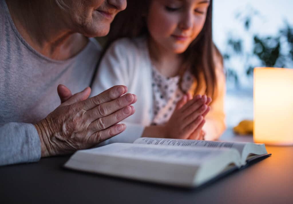 Grandmother and granddaughter praying by lamplight with Bible on table in front of them.