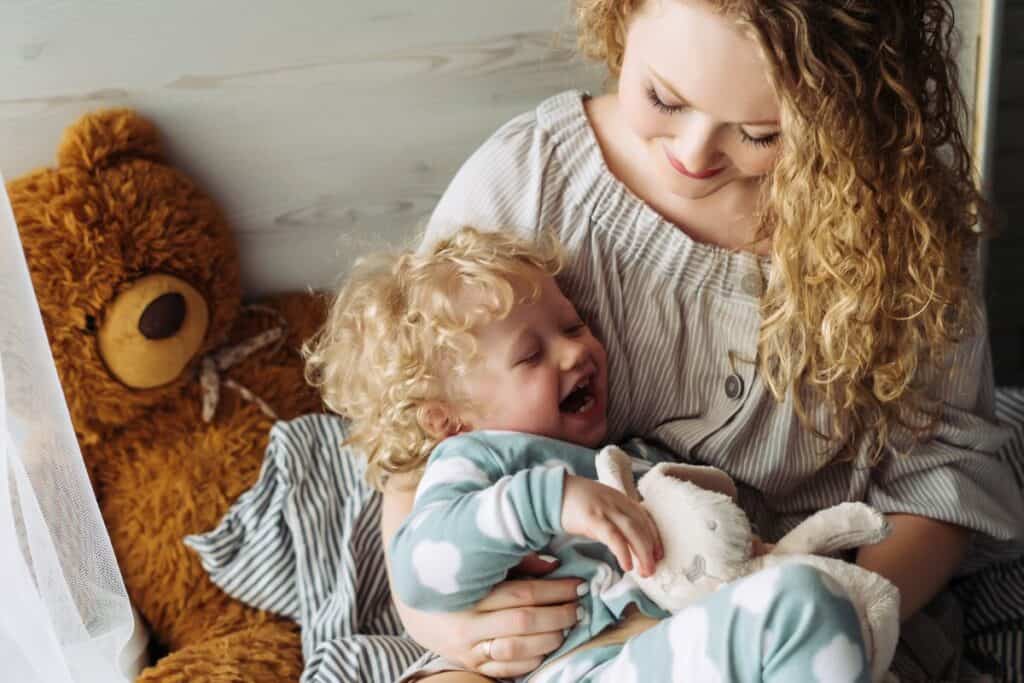 Mom with curly hair smiling down at laughing toddler in her lap.