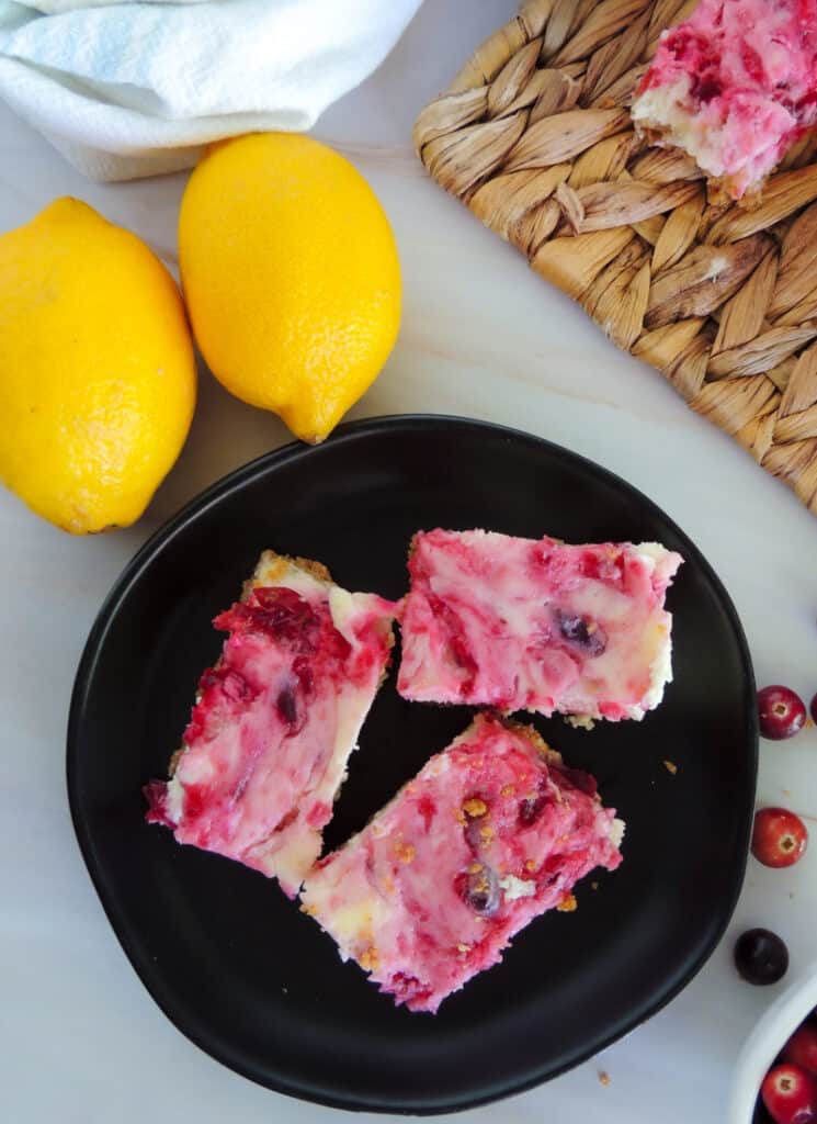 Cranberry cheesecake bars on a black plate with lemons and cranberries on the table nearby.