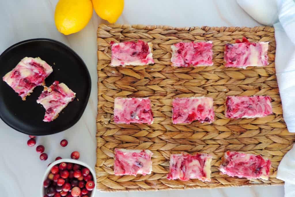 Lemon cranberry cheesecake bars on a woven mat with lemons and cranberries on the table nearby