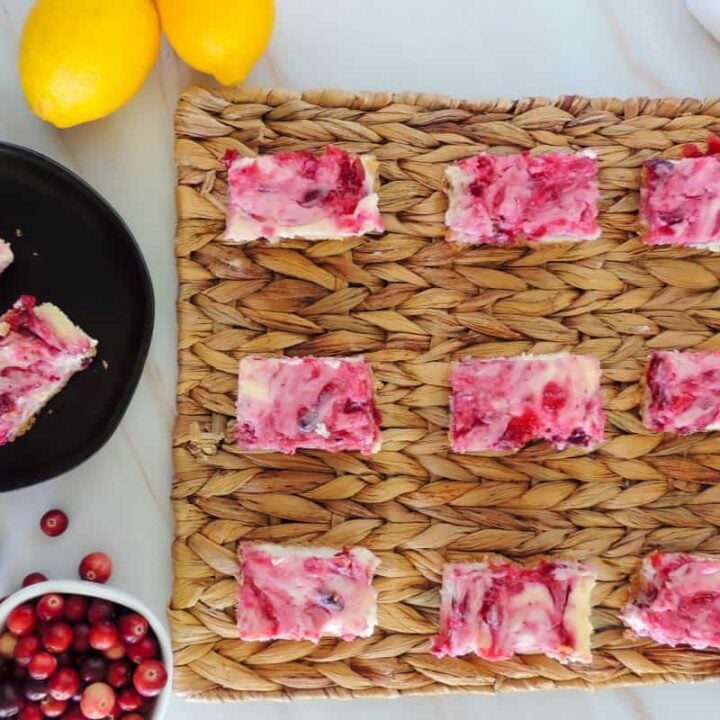 Lemon cranberry cheesecake bars on a woven mat with lemons and cranberries on the table nearby