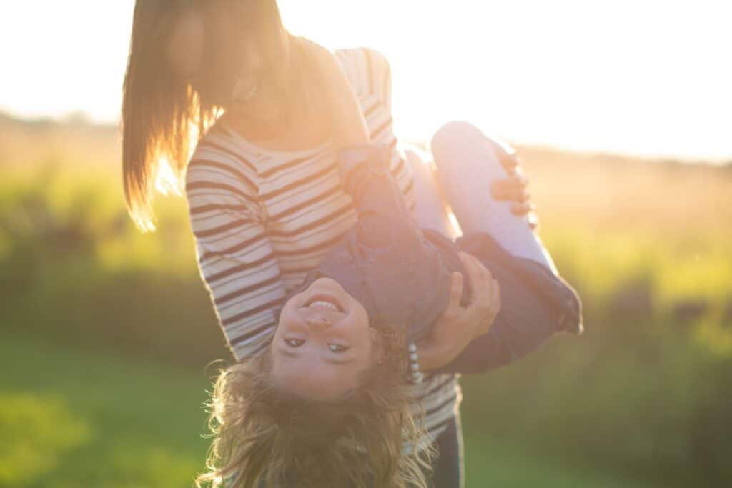 Mom holding toddler girl, spinning her around outside, acting silly together.