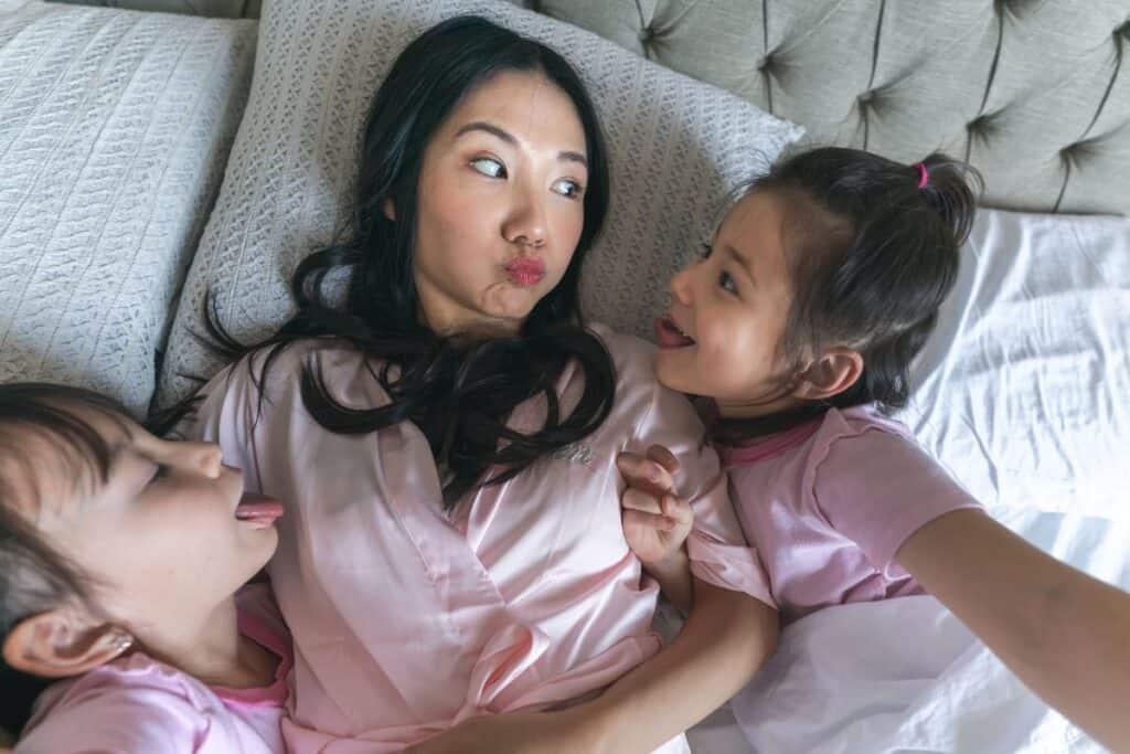 Mom lying on pillows with her two little girls, all making silly faces.