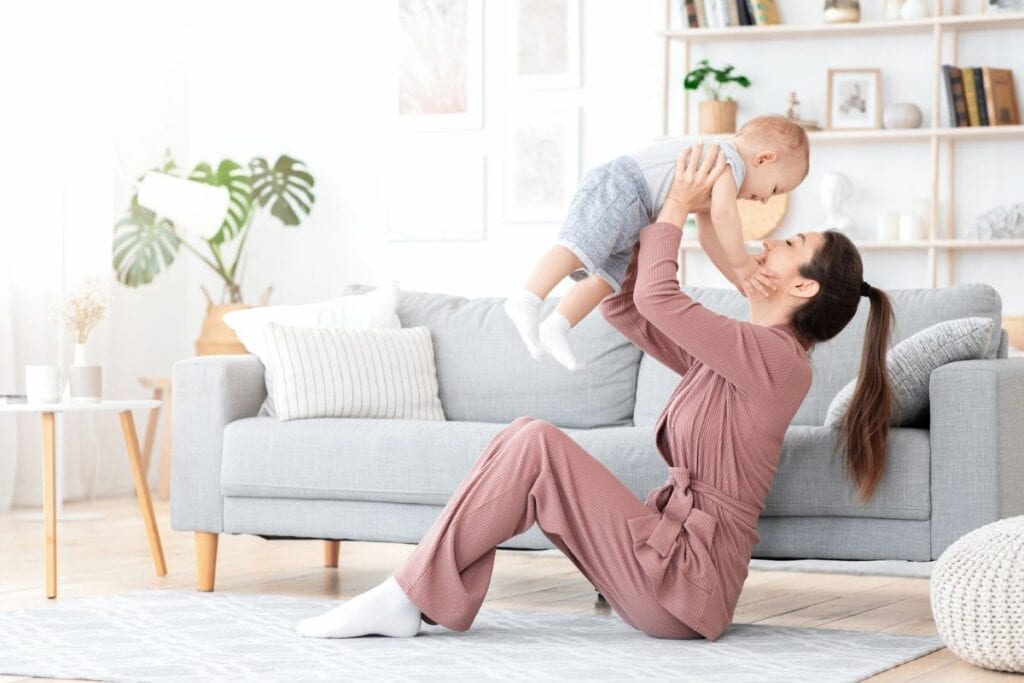 Mom in robe sitting on living room floor, holding baby up in air.