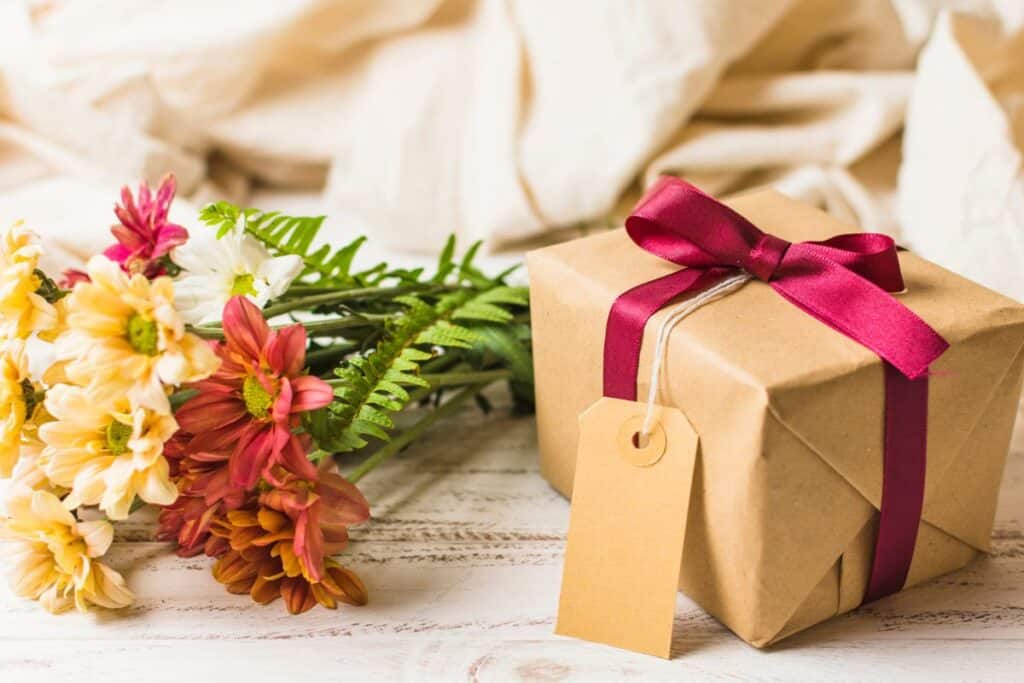 Gift box wrapped in brown paper and red ribbon with colorful spring flowers beside it on wooden table.