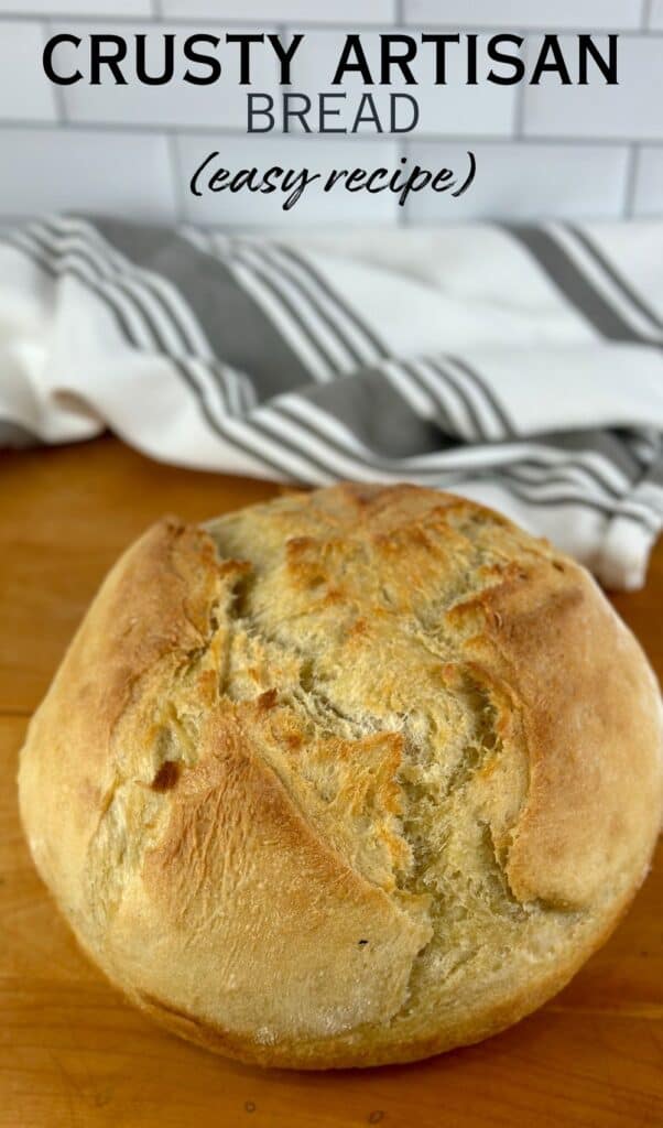 Loaf of crusty round bread loaf on a wooden countertop with striped grey tea towel in background.