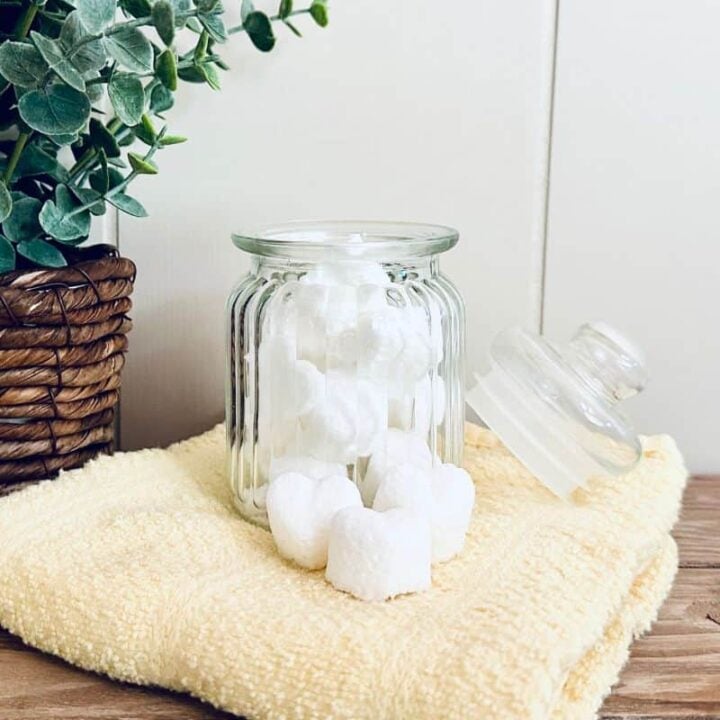 Glass jar of dishwasher tablets with tablets piled on towel nearby and faux plant in background.