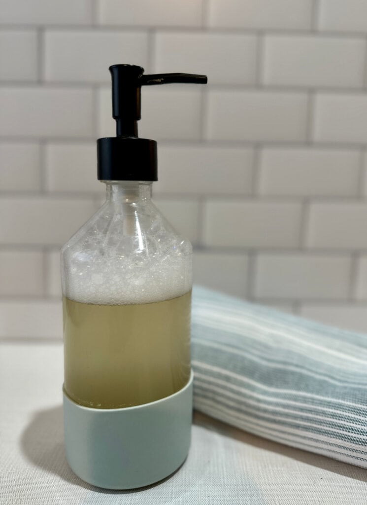 Face wash in glass container with towel in background.