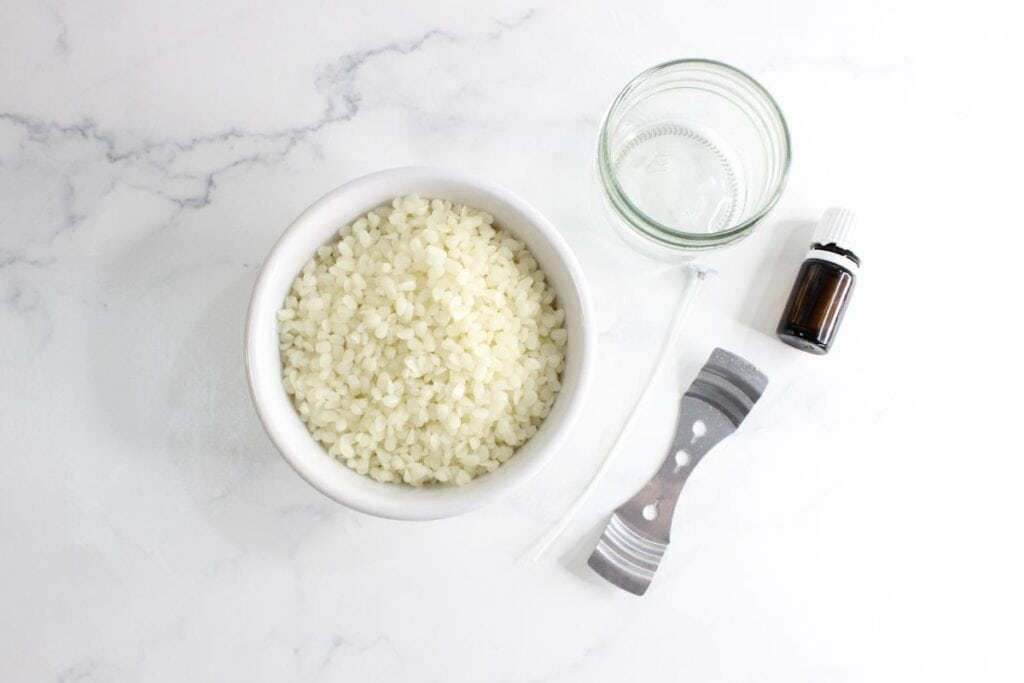 Beeswax pellets, 4 oz glass jar, essential oils jar and wick holder on a marble counter top.
