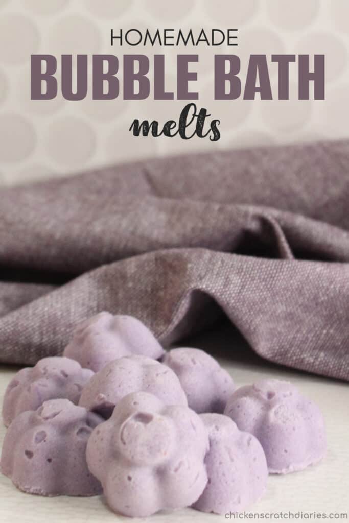 Cluster of lavender colored bubble bath melts in the shape of flowers on tabletop with purple tea towel in background with text "Homemade Bubble Bath Melts"