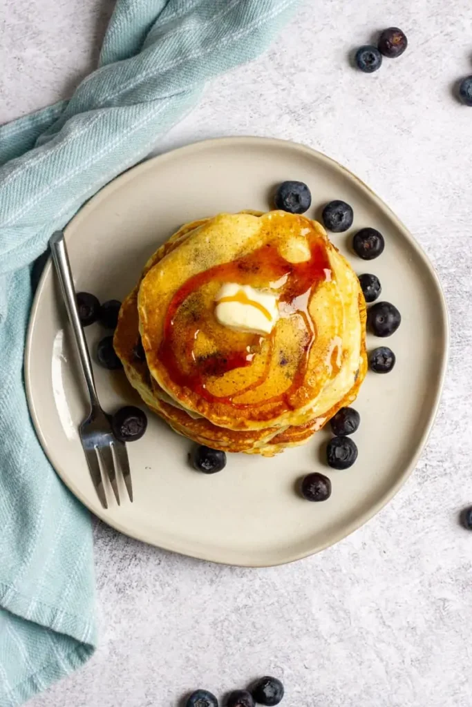 A stack of blueberry pancakes with syrup and topped with blueberries.