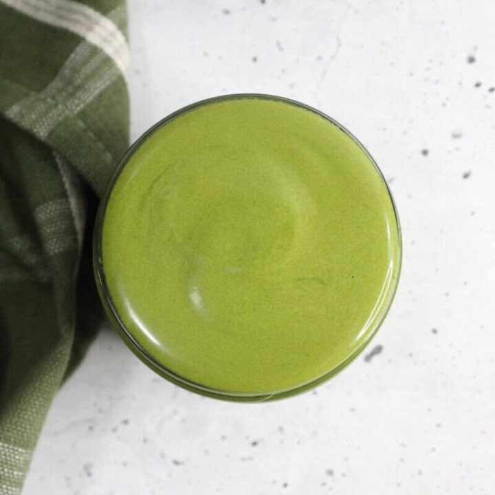 Matcha face cream in small jar next to a green plaid tea towel on countertop.
