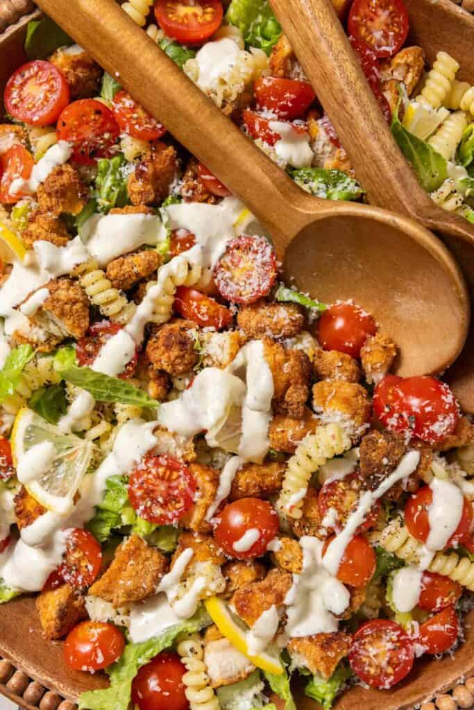 Chicken Caesar pasta salad with two large wooden spoons in the bowl and fresh dressing drizzled on top.