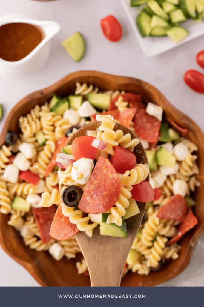 Wooden spoon holding pasta salad with pepperoni above a wooden bowl.