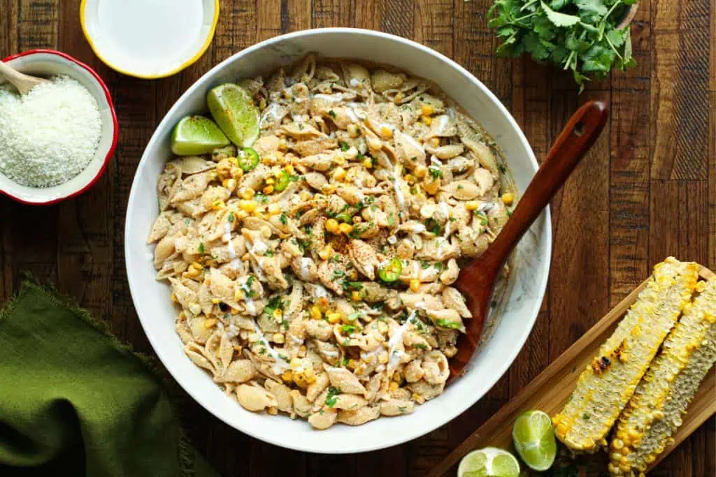 Mexican street corn pasta salad in a large bowl with fresh limes and cut corn on the cob and other ingredients shown nearby.