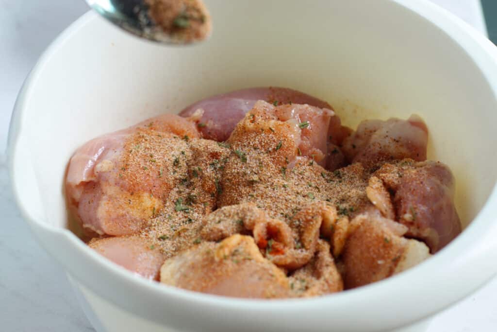 Chicken thighs in large mixing bowl with spices poured on top.