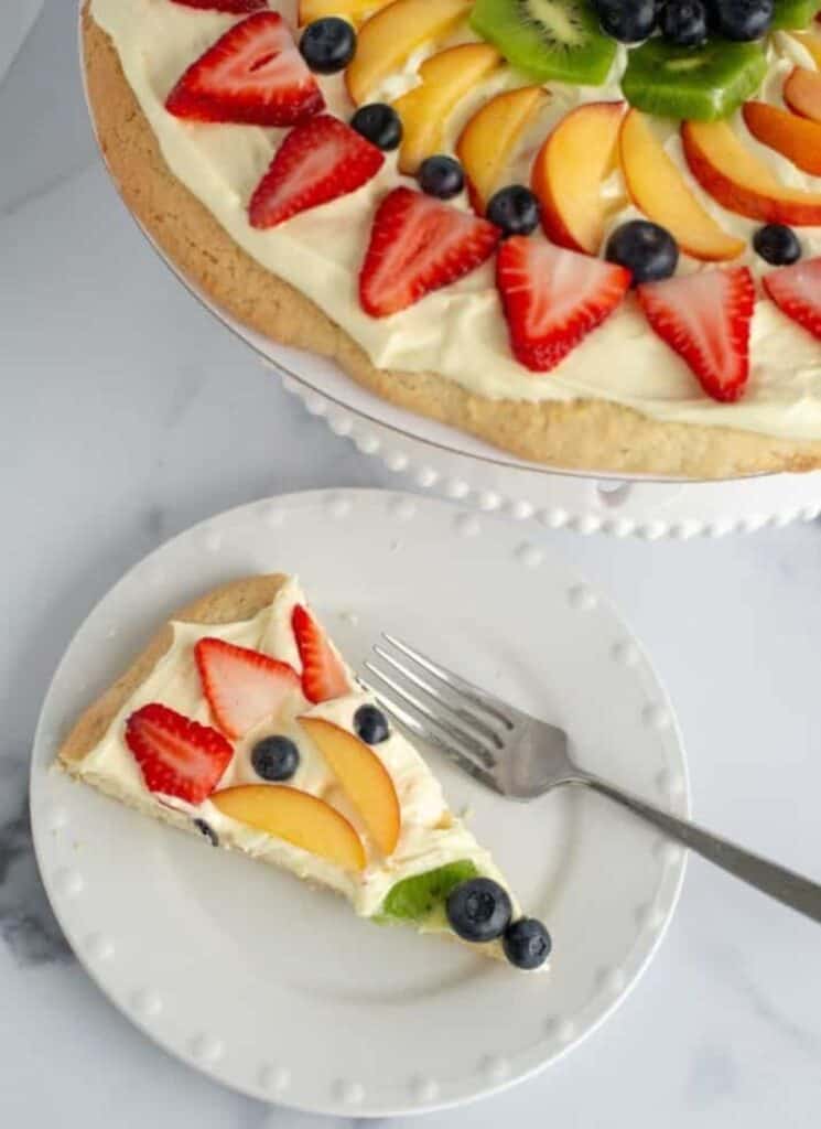 Fruit pizza on cake stand with slice of fruit pizza below on plate.