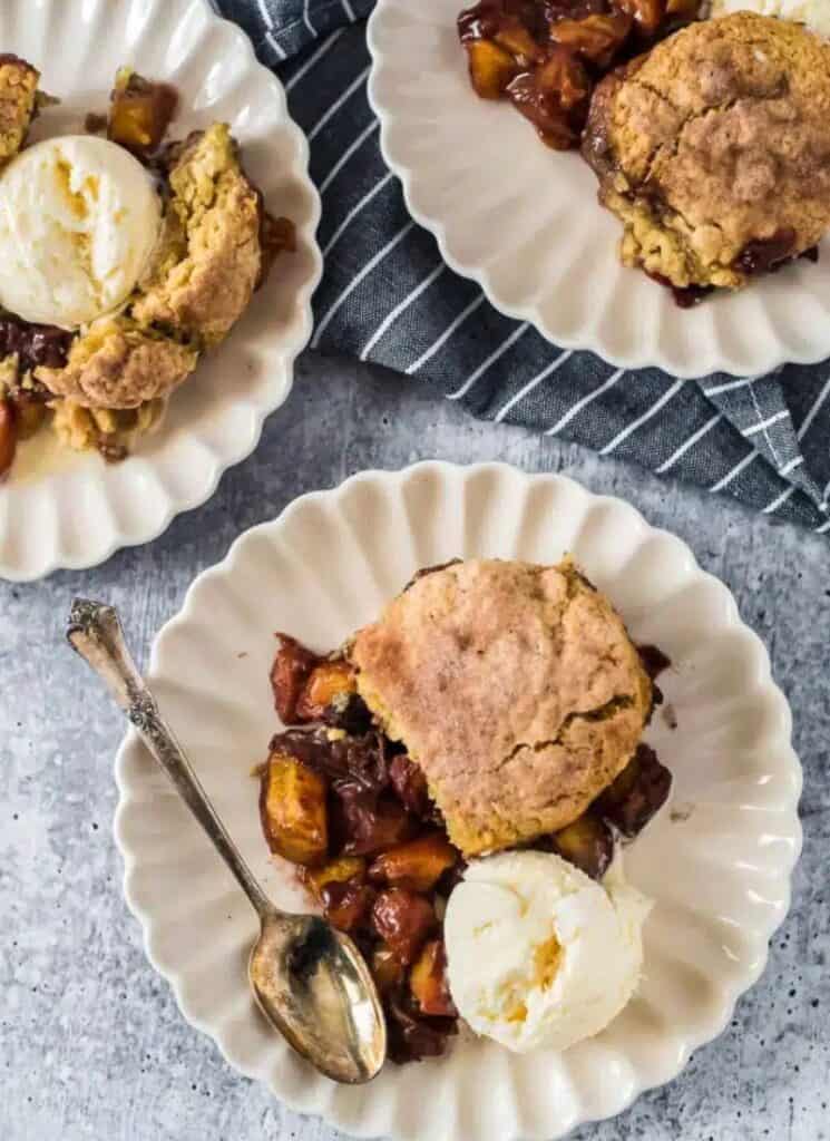 Peach cobbler with sourdough biscuit topping on plate with ice cream and 2 similar plates in background.