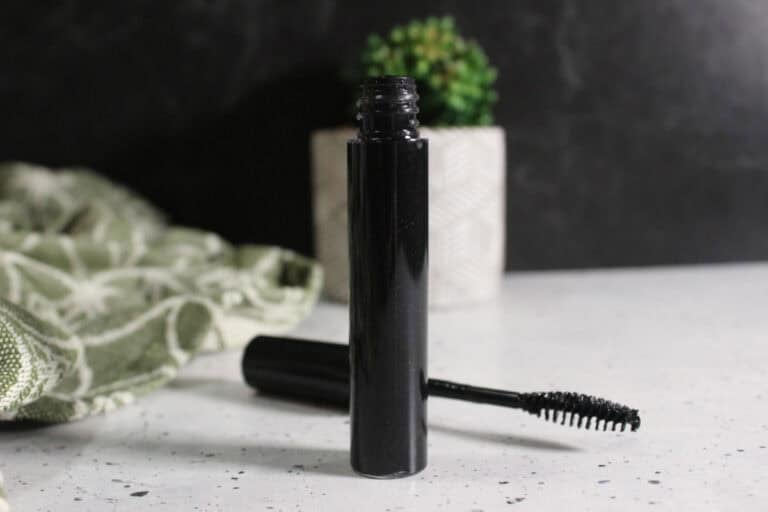 tube of mascara with wand on marble table top beside it and decorate plant nearby.