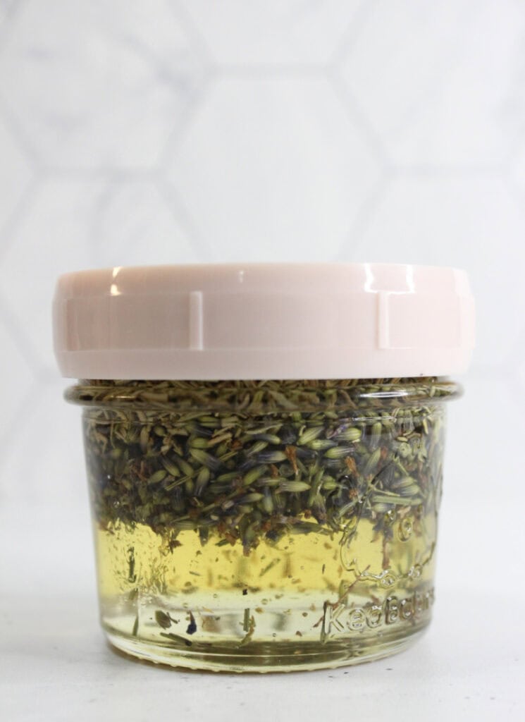 Dried lavender and rosemary leaves suspended in jojoba oil in a small glass jar with a lid.